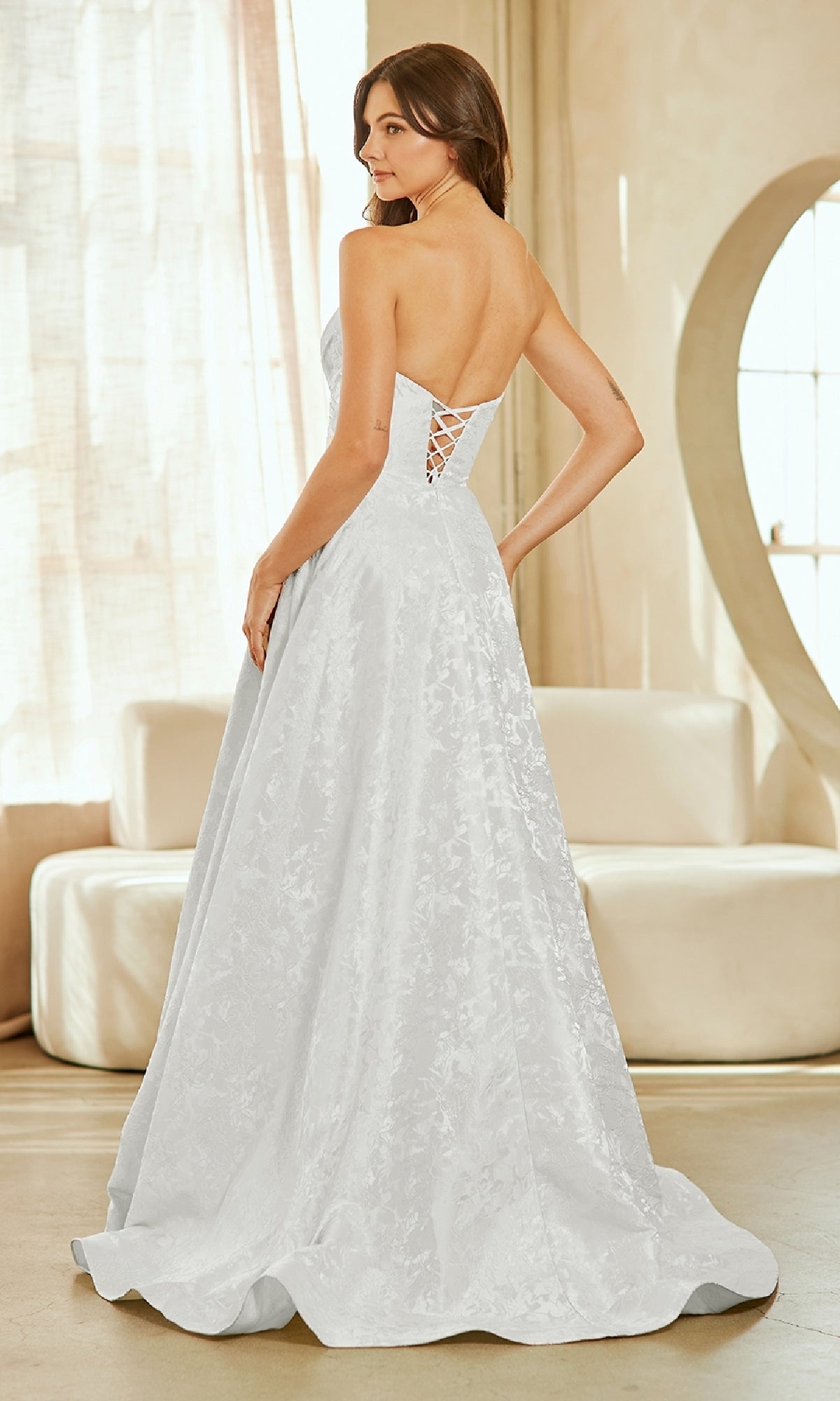 A woman stands indoors wearing the Amelia Couture 8061 Textured Strapless Prom Gown with a lace-up back, looking over her shoulder.